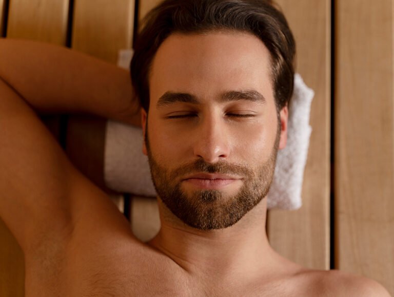 Este hombre está descansando en el banco interior de la sauna. Tiene la cabeza encima de una toalla blanca.