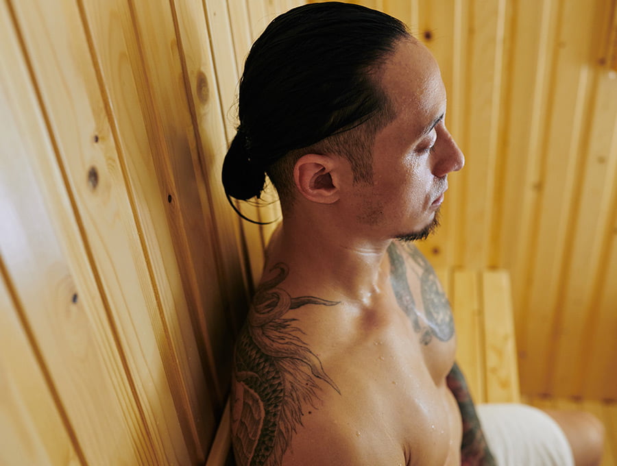 Este hombre está dentro de una sauna con los ojos cerrados. Tiene un tatuaje en la espalda.