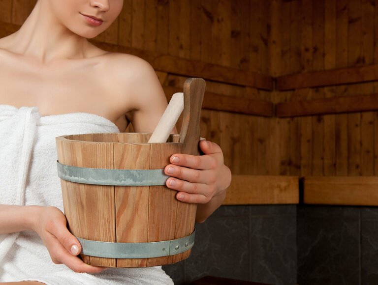 Esta mujer está dentro de la sauna. Tiene el cubo de madera lleno de agu y aroma esencial para poner sobre las rocas calientes.