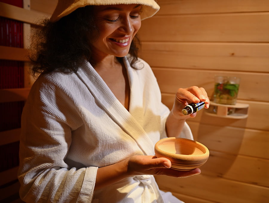 Esta mujer echa nas gotas de aceite esencial de menta en un recipiente dentro de la sauna. Lleva un albornoz.