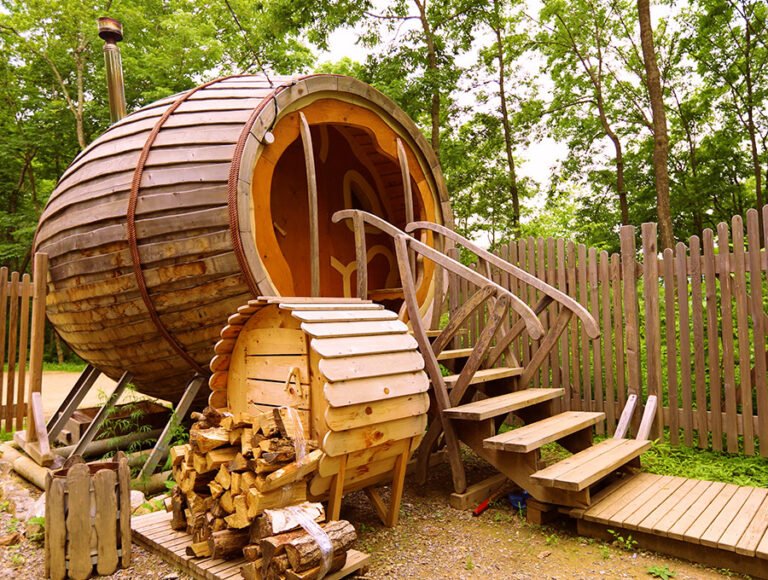 Una sauna con forma de barril colocada en el exterior de un jardín. Todo es de madera.